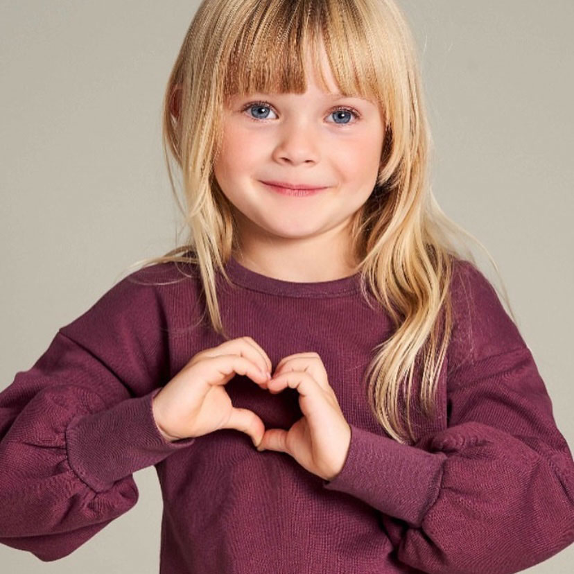 Little girl in a burgundy organic sweatshirt making a heart with her hand